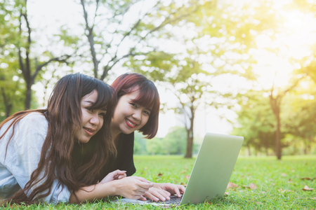 Young asian women lying on grass and using laptop and typing. Girls hands on keyboard. Distance learning concept. Happy hipster young asian women working on laptop in park. Student studying outdoors.の写真素材