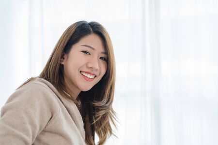 Happy beautiful young Asian woman waking up in morning, sitting on bed, stretching in cozy bedroom, looking through window. Funny asian woman after wake up. Asia woman is stretching and smiling.の写真素材