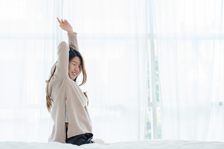 Back view of happy beautiful young Asian woman waking up in morning, sitting on bed, stretching in cozy bedroom, looking through window. Funny woman after wake up. She is stretching and smiling.の写真素材