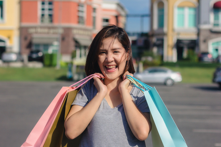 Happy young Asian woman shopping an outdoor market with background of pastel buildings and blue sky. Young asian woman smile with a colorful bag in her hand. Outdoor woman lifestyle shopping concept.の写真素材
