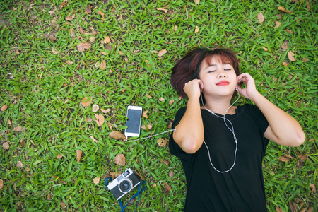 Young Asian woman laying on the green grass listening to music in the park with a chill emotion. Young woman relaxing on the grass with her music playlist. Outdoor activity in the park concept.の写真素材