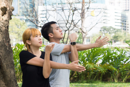 Asian sweet couple warm up their bodies by stretching arms before morning jogging exercise in the park surrounded with nature and warm light sunshine.の写真素材