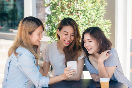 Cheerful asian young women sitting in cafe drinking coffee with friends and talking together. Attractive asian woman enjoying coffee while using smartphone for talking, reading and texting.の写真素材