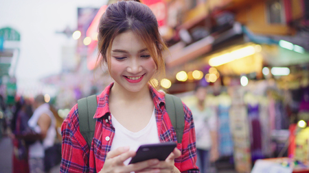Asian woman tourist backpacker smiling and using smartphone traveling alone holidays outdoors on city street in Khao San road at Bangkok Bangkok - Thailand.の写真素材