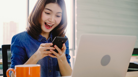 Beautiful young smiling asian woman working on laptop while enjoying using smartphone at office. Asian business woman working document finance in home office. Enjoying time at office.の写真素材