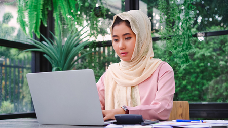 Beautiful young smiling asian muslim woman working on laptop sitting in living room at home. Asian business woman working document finance and calculator in her home office. Enjoying time at home.の写真素材
