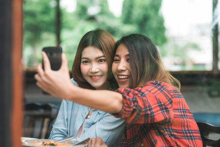 Beautiful happy Asian women lesbian lgbt couple sitting each side eating a plate of Italian seafood spaghetti and french Fries at restaurant or cafe. Female couple using smartphone for selfie.の写真素材