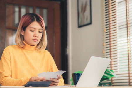 Beautiful young smiling asian woman working laptop on desk in living room at home. Asia business woman writing notebook document finance and calculator in home office. Enjoying time at home concept.の写真素材