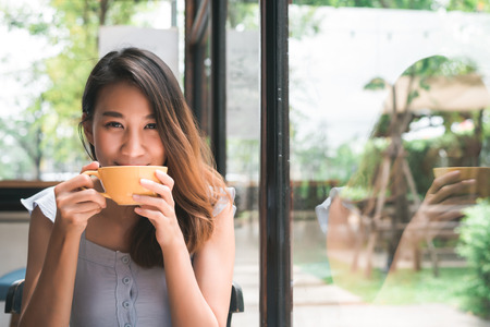 Cheerful asian young woman drinking warm coffee or tea enjoying it while sitting in cafe. Attractive happy asian woman holding a cup of coffee.の写真素材