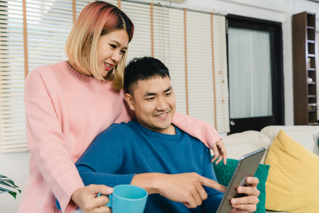 Attractive Asian sweet couple using tablet while lying on the sofa when relax in their living room at home. Husband and his wife using relax and romantic time at home concept.の写真素材