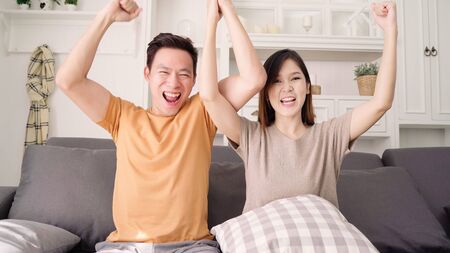 Asian couple cheer football match in front of television living room at home, sweet couple enjoy funny moment while lying on the sofa when relaxed at home. Lifestyle couple relax at home concept.の写真素材