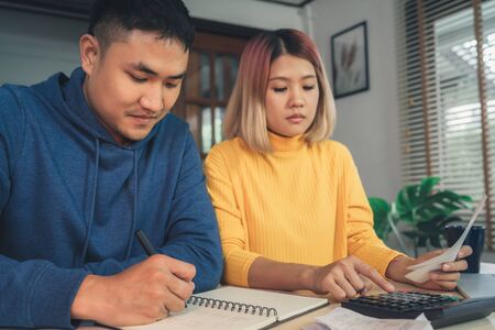 Young asian couple managing finances, reviewing their bank accounts using laptop computer and calculator at modern home. Woman and man doing paperwork together, paying taxes online on notebook pc.の写真素材