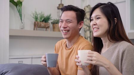 Asian couple watching TV and drinking warm cup of coffee in living room at home, sweet couple enjoy love moment while lying on the sofa when relax at home. Lifestyle couple relax at home concept.の写真素材