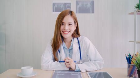 Confident Asian female doctor sitting at office desk and smiling at camera, health care and prevention concept in medical office.の写真素材