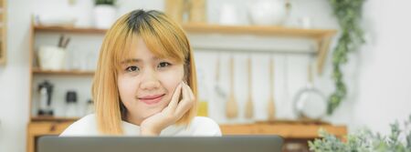 Beautiful young smiling asian woman working on laptop while sitting in a living room at home. Asian business woman working in her home office. Enjoying time at home. Panoramic banner.の写真素材