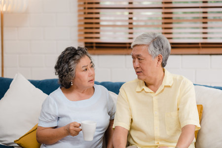 Asian elderly couple drinking warm coffee and talking together in living room at home, couple enjoy...