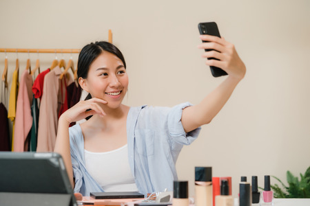 Happy beautiful young Asian woman using smartphone selfie live streaming online to social network. Beauty blogger present beauty cosmetics sitting in front tablet camera for recording video.の写真素材