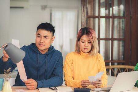 Young stressed asian couple managing finances, reviewing their bank accounts using laptop computer and calculator at modern home. Woman and man doing paperwork together, paying taxes online.の写真素材