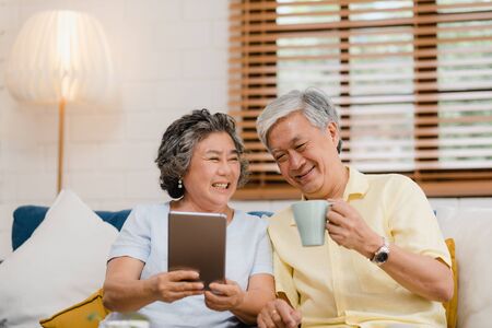 Asian elderly couple using tablet and drinking coffee in living room at home, couple enjoy love moment while lying on sofa when relaxed at home. Enjoying time lifestyle senior family at home concept.の写真素材