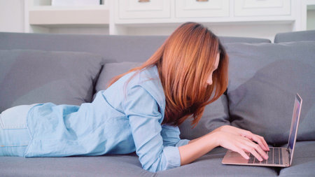 Portrait of beautiful attractive young smiling Asian woman using computer or laptop while lying on the sofa when relax in living room at home. Enjoying time lifestyle women at home concept.の写真素材