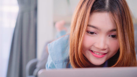 Portrait of beautiful attractive young smiling Asian woman using computer or laptop while lying on the sofa when relax in living room at home. Enjoying time lifestyle women at home concept.の写真素材