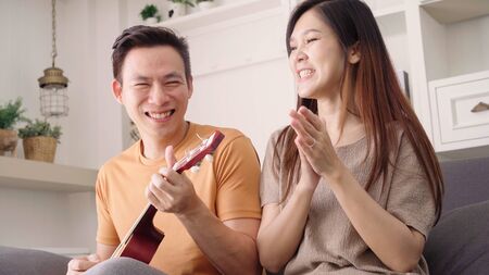 Asian couple playing guitar and singing together in living room at home, sweet couple enjoy love moment while lying on the sofa when relax at home. Lifestyle couple relax at home concept.の写真素材