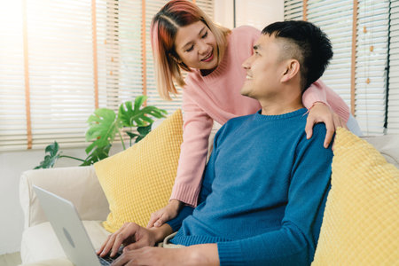 Attractive Asian sweet couple using computer or laptop while lying on the sofa when relax in their...