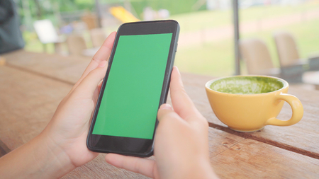 Young asian woman using black mobile phone device with green screen. Asian woman holding smartphone, scrolling pages while sitting in cafe. Chroma key.の写真素材