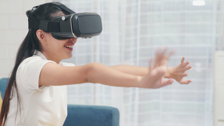 Asian teenager woman using glasses virtual reality simulator playing video games in living room,...