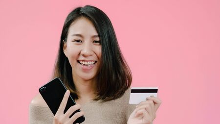 Young Asian woman using smartphone buying online shopping by credit card feeling happy smiling in casual clothing over pink background studio shot. Happy smiling adorable glad woman rejoices success.の写真素材