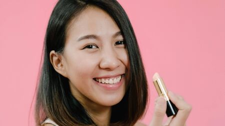 Young beautiful fashionable Asian woman applying lips makeup with cosmetic brush in casual clothing over pink background studio shot. Happy smiling adorable glad woman rejoices success.の写真素材