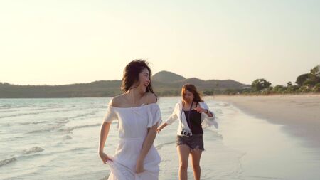 Young Asian lesbian couple running on beach. Beautiful women friends happy relax having fun on beach near sea when sunset in evening. Lifestyle lesbian couple travel on beach concept.の写真素材