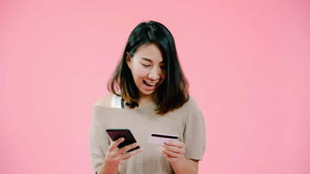 Young Asian woman using smartphone buying online shopping by credit card feeling happy smiling in casual clothing over pink background studio shot. Happy smiling adorable glad woman rejoices success.の写真素材