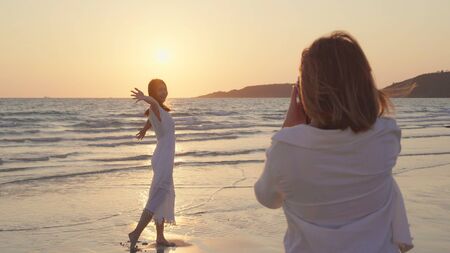 Young Asian lesbian couple using camera taking photo each other near beach. Beautiful women lgbt couple happy romantic moment when sunset in evening. Lifestyle lesbian couple travel on beach concept.の写真素材