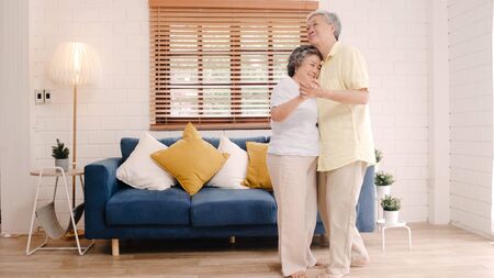 Asian elderly couple dancing together while listen to music in living room at home, sweet couple enjoy love moment while having fun when relaxed at home. Lifestyle senior family relax at home concept.の写真素材