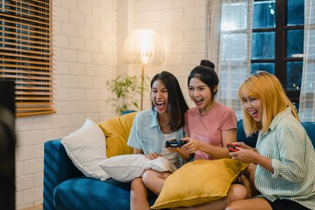 Group of Asian women couple play games at home, female using joystick having funny moment together on sofa in living room in night. Teenager young friend football fan, celebrate holiday concept.の写真素材