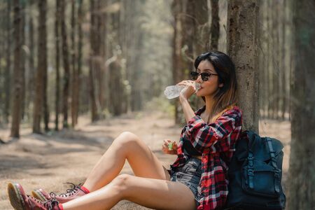 Asian hiker woman trekking in forest. Young happy backpack girl sitting rest drink water from bottle after walking travel nature and adventure trip, climb mountain in summer holidays vacation concept.の写真素材
