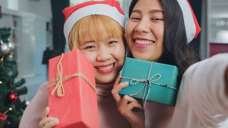 Asian Lesbian couple celebrate Christmas festival. LGBTQ female teen relax happy holding Gift and using smartphone selfie with Christmas tree enjoy xmas winter holidays in living room at home.の写真素材