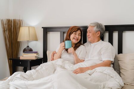 Asian senior couple talking on bed at home. Asian Senior Chinese grandparents, husband and wife happy drink coffee after wake up while lying on bed in bedroom at home in the morning concept.の写真素材
