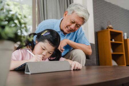 Asian grandfather teach granddaughter drawing and doing homework at home. Senior Chinese, grandpa happy relax with young girl lying on sofa in living room at home concept.の写真素材
