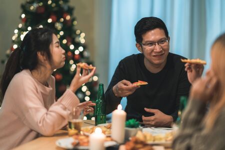 Merry Christmas and Happy New Year! Young friends group having dinner at home. Asian family party with beer and pizza food near christmas tree at house in night. Celebration holiday and togetherness.の写真素材