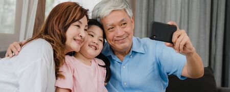 Asian grandparents selfie with granddaughter at home. Senior Chinese happy spend family time relax using mobile phone with young girl kid lying on sofa in living room. Panoramic banner background.の写真素材