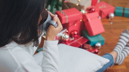 Asian women celebrate Christmas festival. Female teen relax happy having breakfast drink coffee near Christmas tree lying on sofa enjoy xmas winter holidays in living room at home.の写真素材