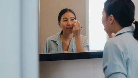 Beautiful Asian woman using eyebrow pencil make up in front mirror, Happy latin female using beauty cosmetics to improve herself ready to working in bathroom at home. Lifestyle women relax at home.の写真素材