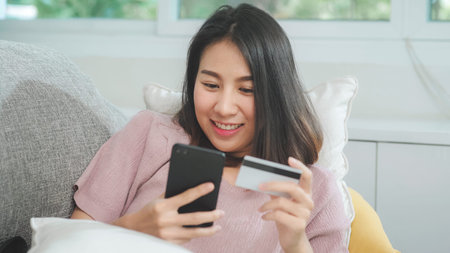 Young smiling Asian woman using smartphone buying online shopping by credit card while lying on sofa...
