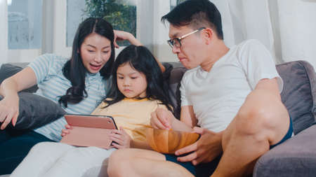 Young Asian family and daughter happy using tablet at home. Japanese mother, father relax with little girl watching movie lying on sofa in living room. Funny parent and lovely child are having fun.の写真素材