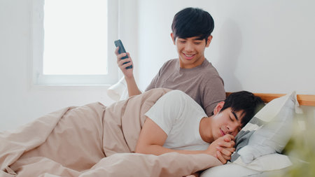 Asian Gay couple using mobile phone at home. Young Asia LGBTQ+ man happy relax rest after wake up,...