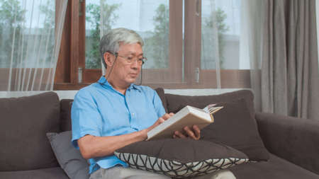 Asian senior men relax at home. Asian Senior Chinese male enjoy rest time read book while lying on sofa in living room at home concept.の写真素材