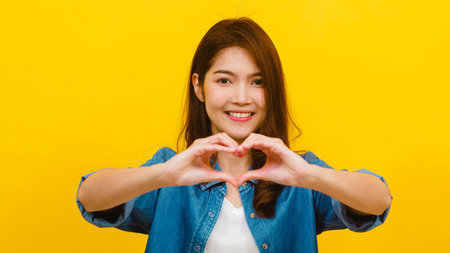 Young Asian lady with positive expression, shows hands gesture in heart shape, dressed in casual...
