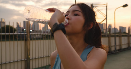 Beautiful young Asia athlete lady exercises drinking water because feel tired after running in urban...